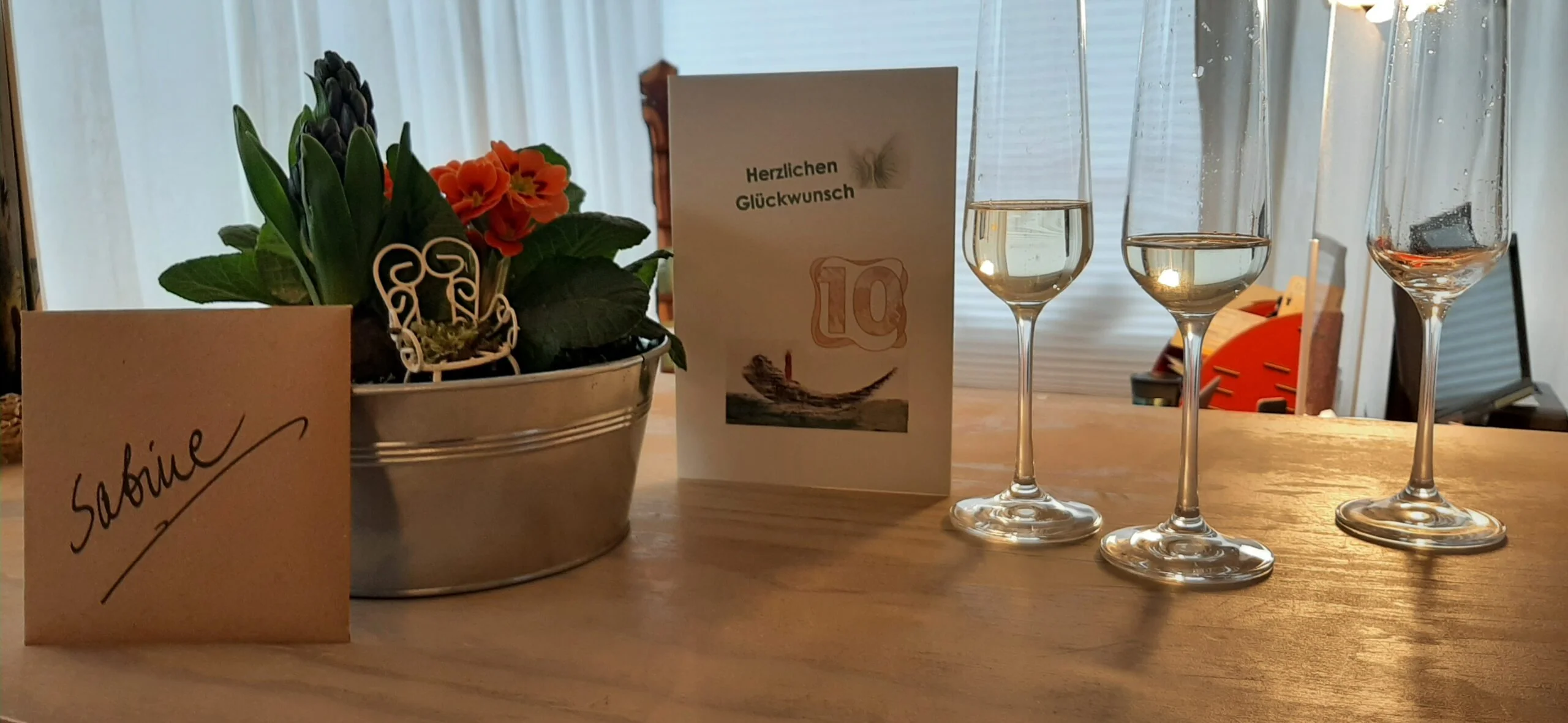 sabine-eller-zehn-jahre-glueckwunsch Glückwunschkarte zum zehnjährigen Jubiläum neben einem Blumengesteck und drei Sektgläsern.