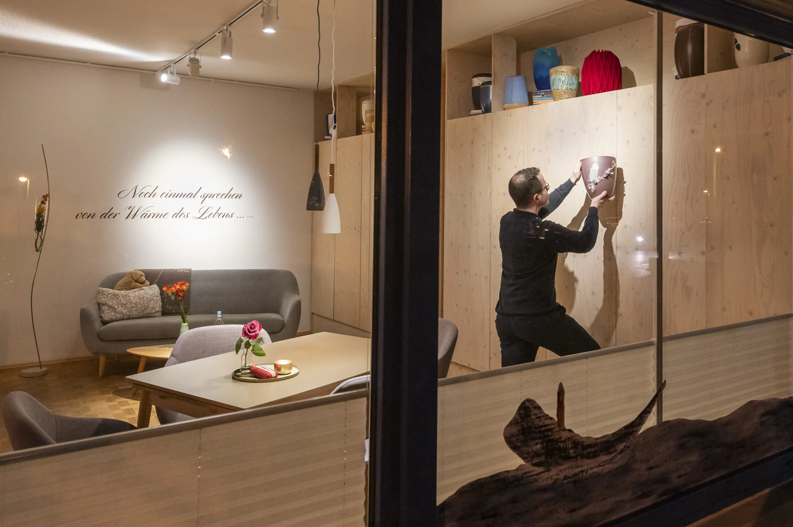 Blick durch ein Schaufenster in einen hellen Raum, in dem eine Person ein Bild an die Wand hängt.