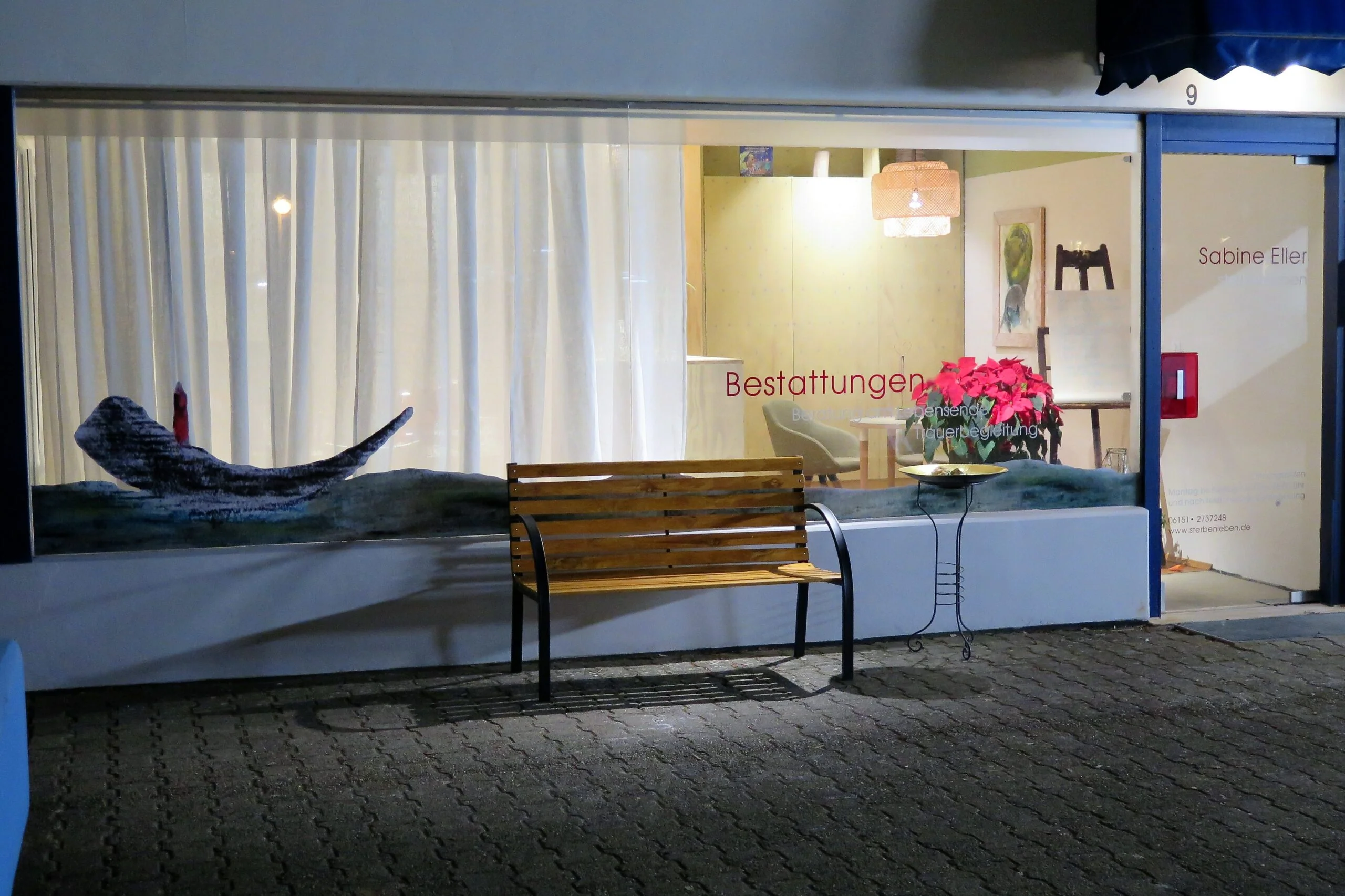 Beleuchtetes Schaufenster von sterbenleben bei Dunkelheit mit einer Sitzbank und Blumen davor.