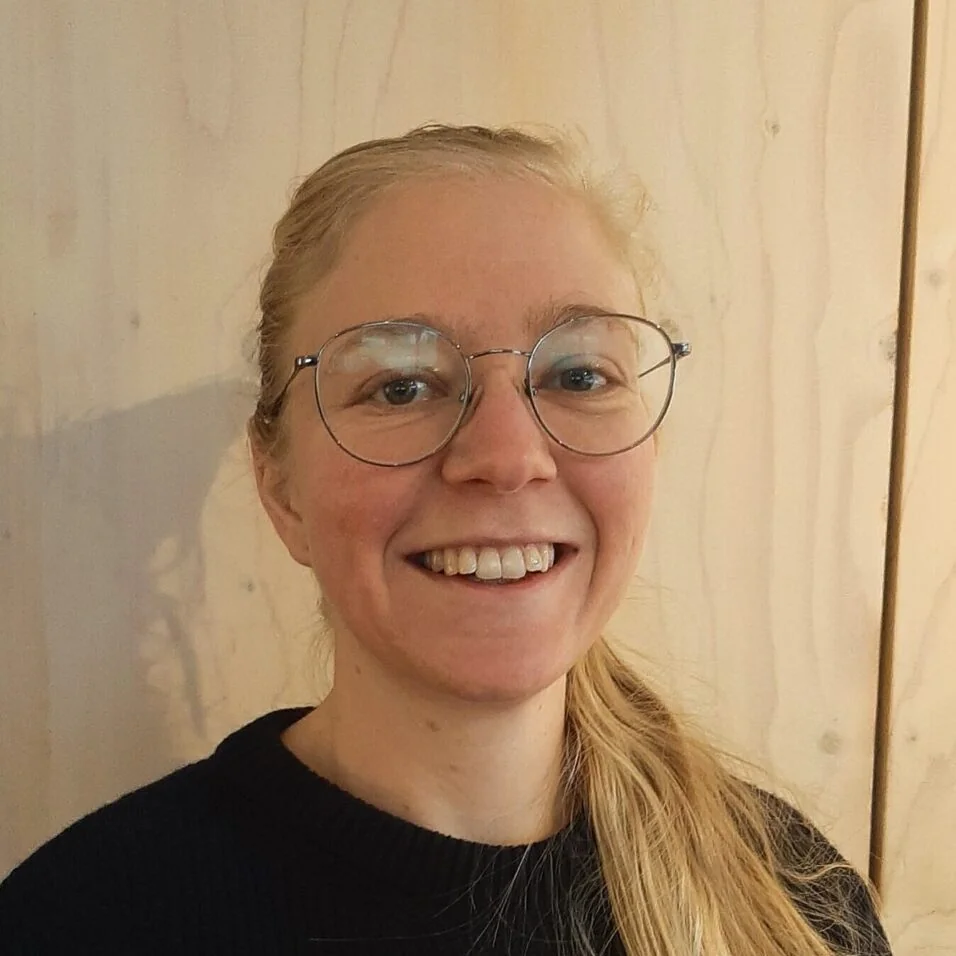 theologinnen-portraet-frau-brille Porträt einer lächelnden Frau mit Brille vor einer hellen Holzwand.